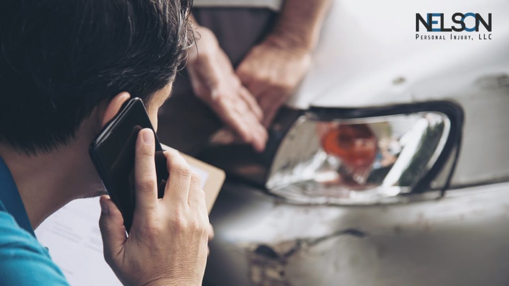 Individual inspecting car damage while talking on phone with Nelson Personal Injury LLC logo